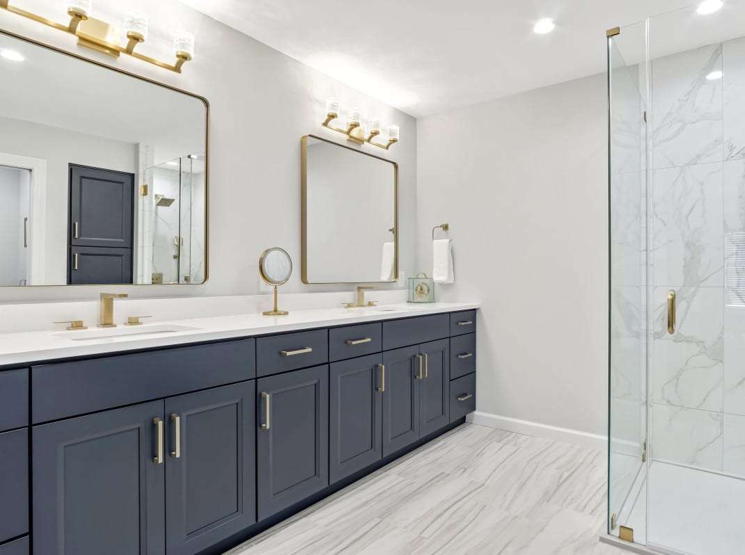 Master bathroom remodel with navy blue custom cabinets, gold framed mirrors and lighting fixtures, porcelain floor and hinged heavy glass shower door.