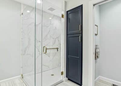 Large master bathroom shower remodel with marble looking porcelain tile with tall navy blue custom cabinet recessed into wall next to shower